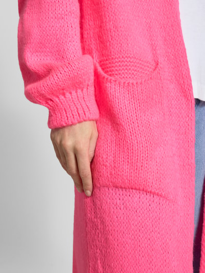 Frauenmodel trägt Longcardigan mit Eingrifftaschen von miss goodlife in leuchtendem Pink, grob gestrickte Baumwoll-Alpaka-Mix Textur, Nahaufnahme Oberkörper.