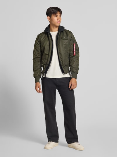 Männermodel trägt Jacke mit Reißverschlusstasche am Ärmel von Alpha Industries in Olivgrün, Regular Fit, gerippte Abschlüsse, Ganzkörperansicht von vorne