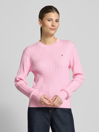 Frauenmodel trägt Slim Fit Pullover aus reiner Baumwolle von Tommy Hilfiger in Rosa mit Zopfmuster, Rundhals und Logo-Detail, Nahaufnahme Oberkörper.