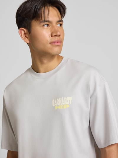 Männermodel trägt Relaxed Fit T-Shirt aus reiner Baumwolle Modell 'Luggage' von Carhartt Work In Progress in Hellgrau, glatte Textur, Nahaufnahme Oberkörper. Herren T-Shirt aus reiner Baumwolle, Relaxed Fit, Label-Detail, Rückenlänge bei Größe M: 78 cm.
