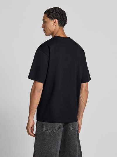 Männermodel trägt Herren T-Shirt mit Statement-Print und Rundhalsausschnitt von Redefined Rebel, Baumwoll-Mix, Relaxed-Fit, Modell Bruno, Rückenlänge 70 cm bei Größe M, schwarze Farbe, Ganzkörperansicht Rückansicht.