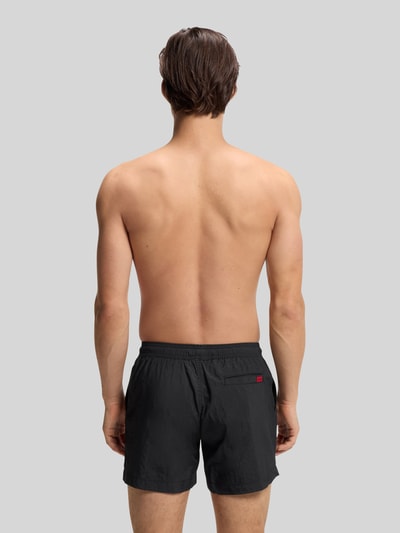 Männermodel trägt Badeshorts mit Label Patch Modell 'DOMINICA' von HUGO in Unifarben, elastischer Bund, Rückentasche, Ganzkörperansicht Rückansicht.