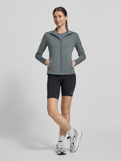 Vrouwenmodel draagt trainingsjack met opstaande kraag en logoprint van Under Armour, materiaalmix met stretch, slim fit, steekzakken, ritssluiting, grijze tinten, ruglengte maat S 59 cm, schouderbreedte 35 cm, borstwijdte 43 cm, mouwlengte 60 cm. Volledige lichaamsvoorzijde.