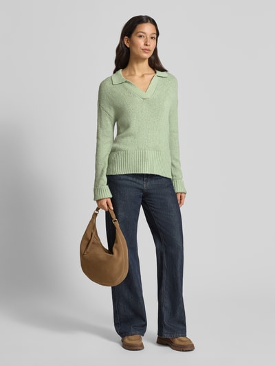 Frauenmodel trägt Strickpullover mit V-Ausschnitt von Fynch-Hatton in Hellgrün, Baumwoll-Mix mit Stretch-Anteil, lockerer Schnitt, Resort-Kragen, gerippte Abschlüsse, kombiniert mit dunkelblauer Hose und brauner Tasche, Ganzkörperansicht von vorne.