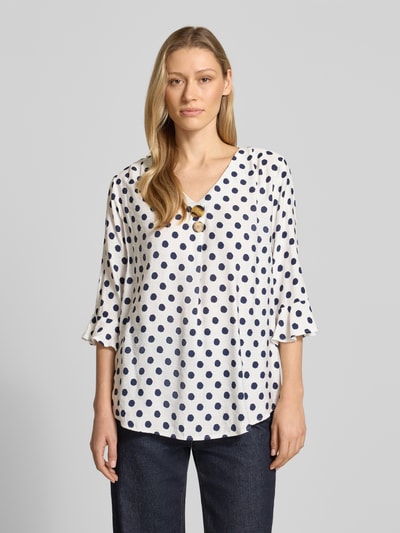Frauenmodel trägt Blusenshirt mit Viskose-Anteil von Apricot, weiß mit dunkelblauen Punkten, Zierknöpfe, 3/4-Arm, Ganzkörperansicht von vorne.