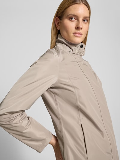 Frauenmodel trägt Regular Fit Mantel mit Logo-Applikation von NAUTGÅRD in Beige, glattes Material, Stehkragen mit Logo-Detail, Nahaufnahme Oberkörper. Rückenlänge 88 cm, Schulterbreite 37 cm, Brustweite 48 cm, Ärmellänge 63 cm, Kapuze mit Tunnelzug, Eingrifftaschen.