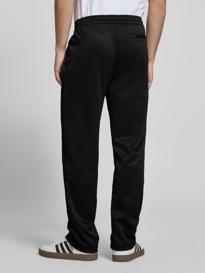 Mężczyzna nosi Trackpants mit Längsnähten Modell 'Motus' od Pegador, czarny materiał z bawełną, krój relaxed fit, szwy wzdłużne, elastyczny pas, długość nogawki 75 cm, widok całej sylwetki z tyłu.