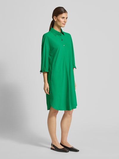 Modelka kobieca ma na sobie zielone, proste Knielanges Hemdblusenkleid mit Knopfleiste od Marc Cain, z kołnierzykiem i rękawem 3/4, widok całej sylwetki z przodu.