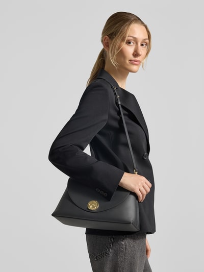 Frauenmodel trägt Handtasche aus echtem Leder Modell 'NIKLA' von Coccinelle in Schwarz mit goldfarbenem Label-Detail, Nahaufnahme Oberkörper. Echtes Leder, abnehmbarer Schulterriemen, Überschlag mit Hauptfach, Innentasche mit Reißverschluss, Maße: 25x17x11 cm, Schulterriemen 88 cm.