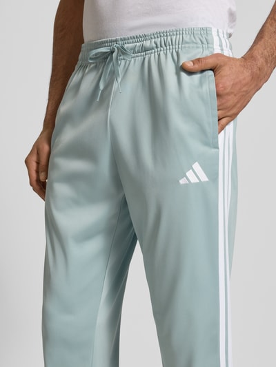 Mężczyzna nosi Slim Fit Sweatpants mit Logo-Stitching von adidas Sportswear, jasnoniebieskie, wpuszczane kieszenie, elastyczny pas z tunelem, krój slim fit, wyhaftowane logo, elastyczne zakończenia nogawek, długość wewnętrzna nogawki 76 cm w rozmiarze M, zbliżenie na górną część spodni z przodu.