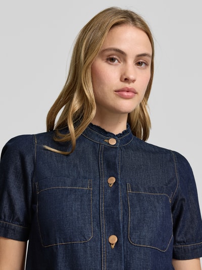 Modelka kobieca nosi Regular Fit Jeansbluse mit Brusttaschen von More & More, czysta bawełna, krój regular fit, stebnowane kieszenie, zapięcie na guziki, stójka, długość tyłu 52 cm, długość rękawa 25 cm, ciemny denim, tekstura gładka, zbliżenie na górną część ciała