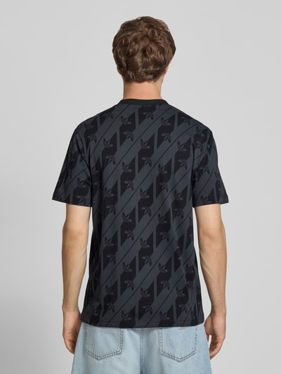 Männermodel trägt T-Shirt aus reiner Baumwolle Modell 'MONOGRAMM AOP' von adidas Originals, dunkle Farben, diagonales Muster, Ganzkörperansicht Rückansicht.