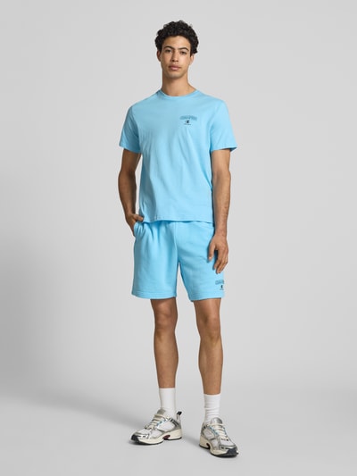 Männermodel trägt T-Shirt mit Logo und Rundhalsausschnitt von CHAMPION in Hellblau, Regular Fit, reine Baumwolle, Ganzkörperansicht von vorne.