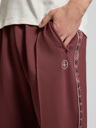 Männermodel trägt Trainingshose mit Biese, aufgesetztem Band und Logo-Bestickung von REVIEW in Bordeauxrot, Nahaufnahme Oberschenkel.