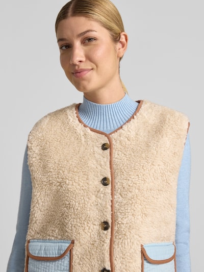 Frauenmodel trägt Weste in Teddy-Optik Modell 'Jace' von LOLLYS LAUNDRY, beige mit braunen Details, blaue Pattentaschen, Nahaufnahme Oberkörper. Gerade Passform, Rundhalsausschnitt, Knopfleiste, Aufgesetzte Pattentaschen.