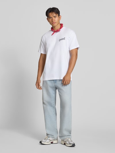 Männermodel trägt Regular Fit T-Shirt aus reiner Baumwolle Modell 'Pup' von Carhartt Work In Progress, weiß, Label-Print, Ganzkörperansicht von vorne. Herren T-Shirt, Rückseitiger Motiv-Print, Rückenlänge bei Größe M: 77 cm.