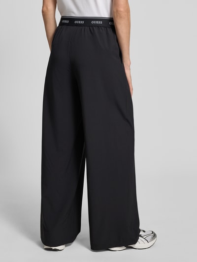Frauenmodel trägt schwarze Wide Fit Pants mit elastischem Bund Modell 'SELENE' von Guess Activewear, Material-Mix, Ganzkörperansicht Rückansicht.