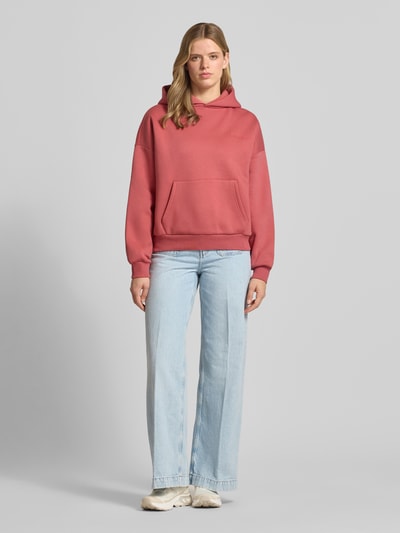 Frauenmodel trägt Hoodie mit Logo und Känguru-Tasche von REVIEW, Damen, Baumwoll-Mix, Relaxed-Fit, rosafarbene Oberfläche mit strukturierter Textur, Ganzkörperansicht von vorne.