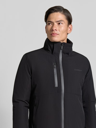 Mężczyzna nosi Regular Fit Parka mit Logo-Print od Lindbergh, elastyczna mieszanka materiałów, czarny kolor, zamek błyskawiczny, napoleońska kieszeń, nadruk z logo, kaptur z tunelem, długość tyłu w rozmiarze M: 90 cm, szerokość ramion w rozmiarze M: 50 cm, szerokość klatki piersiowej w rozmiarze M: 58 cm, długość rękawów w rozmiarze M: 65 cm, zbliżenie górnej części ciała z przodu.