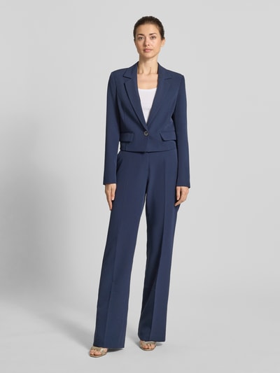 Frauenmodel trägt Cropped Blazer mit Reverskragen Modell 'Leila' von Guess aus Elastischer-Material-Mix, Farbe Dunkelblau, ganzkörperansicht von vorne, Pattentaschen, Knopfverschluss, Rückenlänge 47 cm, Brustweite 42 cm, Ärmellänge 64 cm.