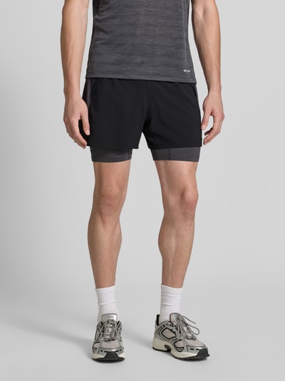 Męski model nosi Regular Fit Shorts mit integrierten Tights Model 'Road' von ASICS TIGER, czarny, detal z logo, krój regular fit, elastyczny pas, Szerokość w rozmiarze M: 38 cm, Długość wewnętrzna nogawki w rozmiarze M: 12 cm, Ganzkörperansicht von vorne.