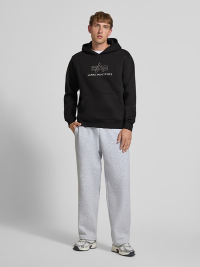 Männermodel trägt Regular Fit Hoodie aus reiner Baumwolle mit Label-Detail von Alpha Industries, Frontansicht Ganzkörperansicht, schwarze Farbe, Känguru-Tasche, elastische Abschlüsse, Rückenlänge 70 cm, Ärmellänge 64 cm.