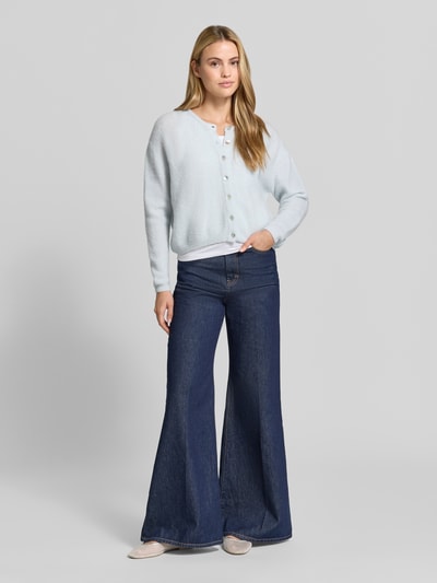 Frauenmodel trägt Relaxed Fit Strickjacke aus Woll-Mix mit Leinen-Anteil von American Vintage in hellem Blau, gerippte Bündchen, durchgehende Knopfleiste, Langarm, kombiniert mit dunkelblauer Schlagjeans, Ganzkörperansicht von vorne.