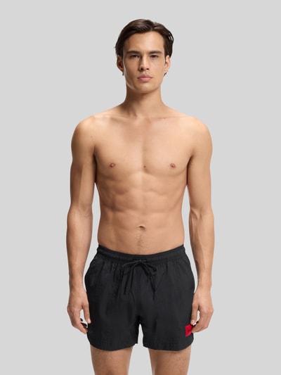 Männermodel trägt schwarze Badeshorts mit Label Patch Modell 'DOMINICA' von HUGO, unifarben, elastischer Bund, Ganzkörperansicht von vorne.