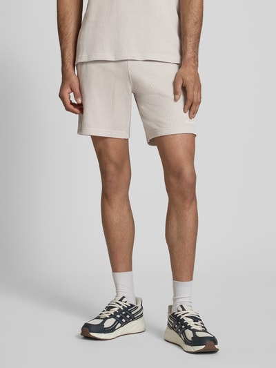 Mężczyznamodel nosi Shorts mit Strukturmuster und elastischem Bund von CHAMPION, jasny beż, krój regular fit, bawełniany miks, strukturalna faktura, label-stitching, szerokość w rozmiarze M: 40 cm, długość nogawki wewnętrznej: 19 cm, całopostaciowy widok z przodu.