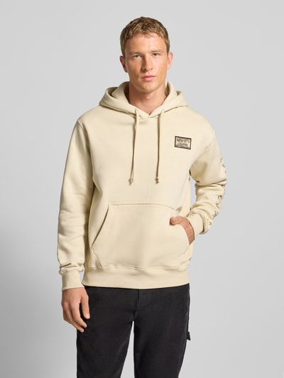 Męski model nosi kremowy Hoodie mit Label-Stitching Modell 'Camo Sleeve' od Alpha Industries, regular fit, przód, ujęcie całej sylwetki.