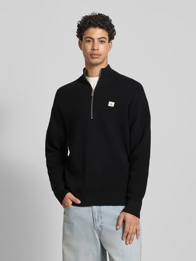 Männermodel trägt Loose Fit Troyer mit gerippten Abschlüssen von Calvin Klein Jeans in Schwarz, kurzer Reißverschluss, Ganzkörperansicht von vorne.
