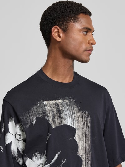 Männermodel trägt Comfort Fit T-Shirt mit Motiv-Print von Armani Exchange in Schwarz mit Allover-Look und Rundhalsausschnitt, Nahaufnahme Oberkörper. Rückenlänge bei Größe M: 76 cm, Herren T-Shirt.