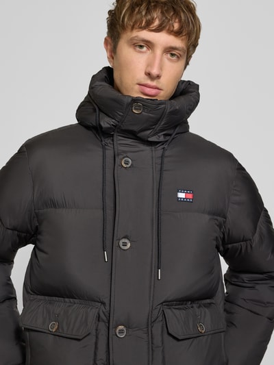 Männermodel trägt Regular Fit Pufferjacke mit Kapuze von Tommy Jeans in Schwarz, Logo-Patch, Steppoptik, Nahaufnahme Oberkörper.