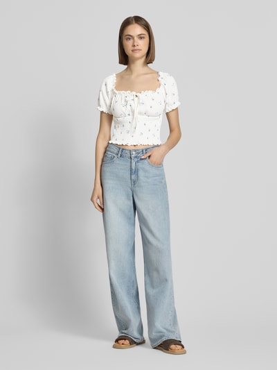Frauenmodel trägt Slim Fit Blusentop mit Allover-Muster Modell 'HILLARY' von Only, floral, cropped, Henley-Ausschnitt, Kurzarm, Raffungen, Weiß mit blauem Muster, leichte Struktur, Ganzkörperansicht von vorne.