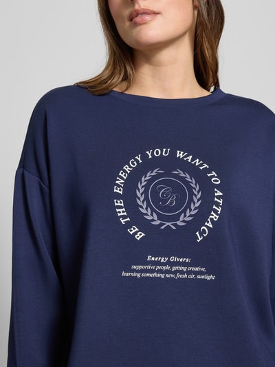 Frauenmodel trägt Sweatshirt mit Bändel und Frontprint von Christian Berg Woman, Oversized, Material-Mix mit Modal-Anteil, Rundhalsausschnitt, navyblau, weißer Print auf Vorderseite, Nahaufnahme Oberkörper.