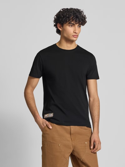 Männermodel trägt T-Shirt mit Label-Patch Modell 'Camo' von Alpha Industries, schwarze Reine gewachste Baumwolle, Gerader-Schnitt, Ganzkörperansicht von vorne.