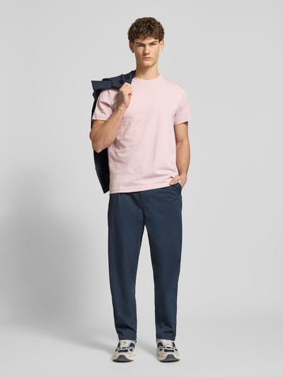 Mężczyzna model nosi T-Shirt aus Baumwolle od G-Star Raw, męski t-shirt, bawełna, prosty krój, okrągły dekolt, wyhaftowane logo, długość tyłu w rozmiarze M: 71 cm, jasnoróżowy, gładka tekstura, ciemnobłękitne spodnie, sneakersy, jasne tło, całkowity widok z przodu.