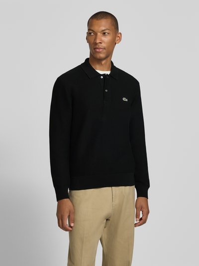 Męski model nosi czarną koszulkę polo Regular Fit Poloshirt aus reiner Baumwolle od Lacoste, beżowe spodnie, całkowity widok z przodu.