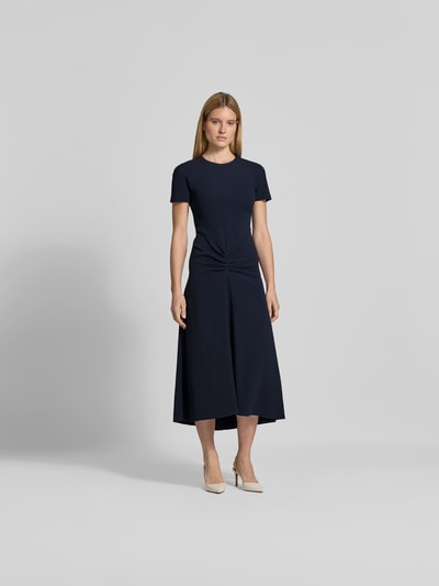 Frauenmodel trägt Midikleid aus Viskose-Mix von Victoria Beckham, tailliert mit Raffungen, kurzärmelig, dunkelblau, Ganzkörperansicht von vorne.