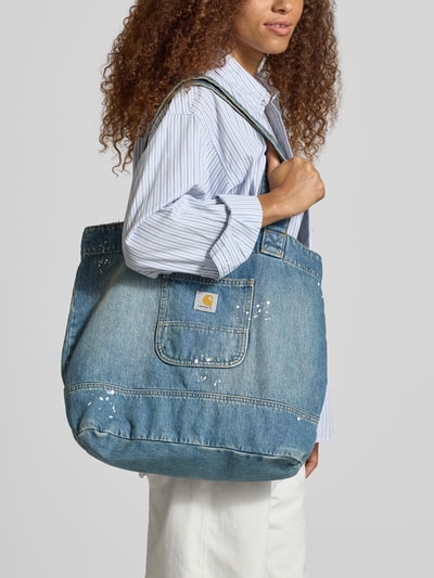 Frauenmodel trägt Tote Bag aus reiner Baumwolle in Denim-Optik von Carhartt Work In Progress, Hauptfach mit Druckknopfverschluss, Logo-Patch, breite Trageriemen, dezente Used-Effekte, Nahaufnahme Oberkörper.
