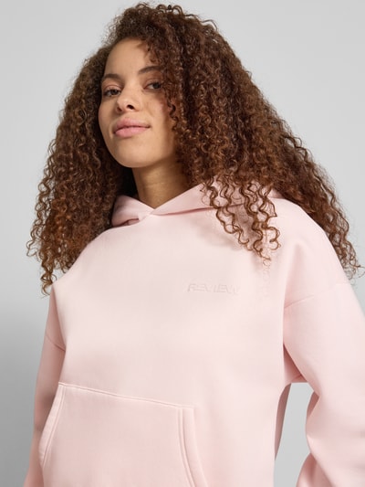 Vrouwenmodel draagt roze Hoodie met Logo-Print en kangoeroezak van REVIEW van katoenmix, loose fit, capuchon, logoprint, Nahaufnahme Oberkörper.