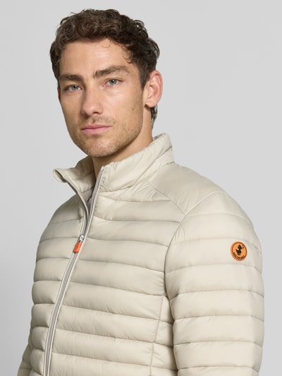 Männermodel trägt Steppjacke mit Zweiwege-Reißverschluss Modell 'ALEXANDER' von Save The Duck in Beige, leichte Steppstruktur, Nahaufnahme Oberkörper. Slim Fit, Stehkragen, Reißverschlusstaschen, Größe M 67 cm Rückenlänge, 47 cm Schulterbreite, 57 cm Brustweite, 68 cm Ärmellänge.