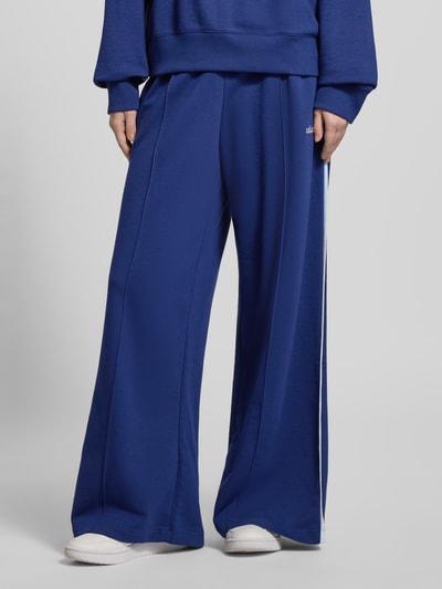 Kobiece model nosi Sweatpants mit elastischem Bund od adidas Sportswear, bawełniany miks z wiskozą, luźny krój, elastyczny pas, label-stitching, długość nogawki wewnętrznej w rozmiarze S: 77 cm, kolor niebieski, rozcięcia po bokach, zbliżenie na dolną część ciała.