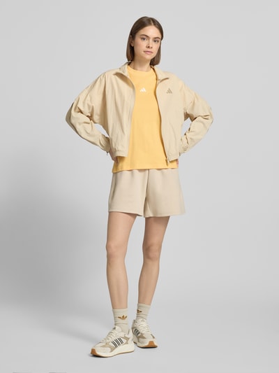 Vrouwenmodel draagt Jacke mit Stehkragen van adidas Sportswear in regular fit, lichtbeige tinten, elastische boorden, labelprint, ritssluiting, vooraanzicht volledig lichaamsfoto. Lengte rug maat S: 55 cm, schouderbreedte: 66 cm, borstwijdte: 55 cm, mouwlengte: 48 cm.