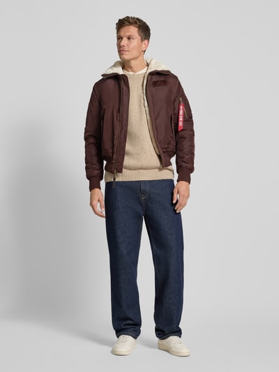 Männermodel trägt braune Bomberjacke mit Reißverschluss und Label-Patch von Alpha Industries mit Kunstfellkragen, Ganzkörperansicht von vorne. Regular Fit, kombiniert mit beiger Strickware und blauen Jeans, passende Größenangaben im Kontext.