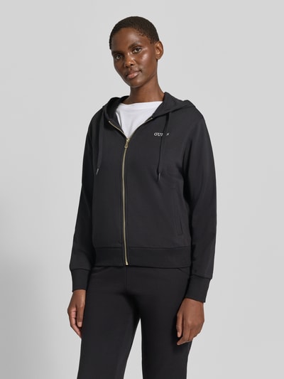 Vrouwenmodel draagt zwarte sweatjack met capuchon en ritssluiting, katoenmix, regular fit, model 'SOFIA' van GUESS, labelstitching. Ganzkörperansicht von voren.