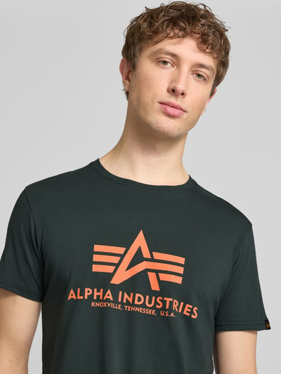 Męski model nosi t-shirt z detalem z logo model 'BASIC' od Alpha Industries, bawełna, prosty krój, okrągły dekolt, długość tyłu 69 cm w rozmiarze M, zbliżenie na górę ciała