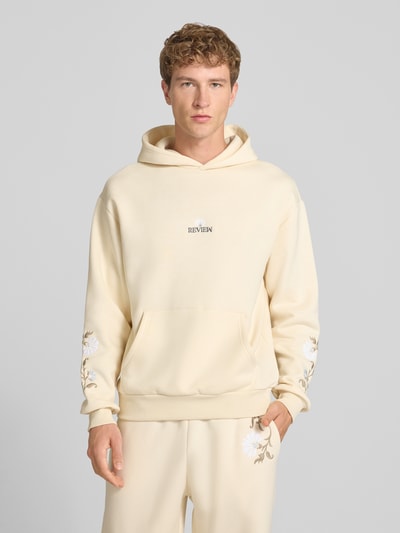 Männermodel trägt Relaxed Fit Hoodie mit Känguru-Tasche von REVIEW in Creme mit floraler Stickerei, Baumwoll-Mix, Nahaufnahme Oberkörper.