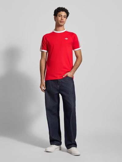 Männermodel trägt T-Shirt mit Label-Stitching von adidas Originals in Rot mit weißen Details, Reine Baumwolle, Regular-Fit, Ganzkörperansicht von vorne.