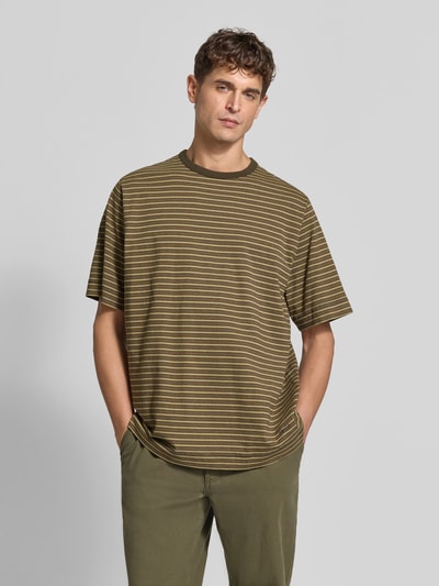 Männermodel trägt Relaxed Fit T-Shirt mit Label-Stitching von Forét, Streifenmuster in Oliv und Weiß, Rundhalsausschnitt, Halbarm, reine Baumwolle, Rückenlänge 73 cm bei Größe M, Ganzkörperansicht von vorne.